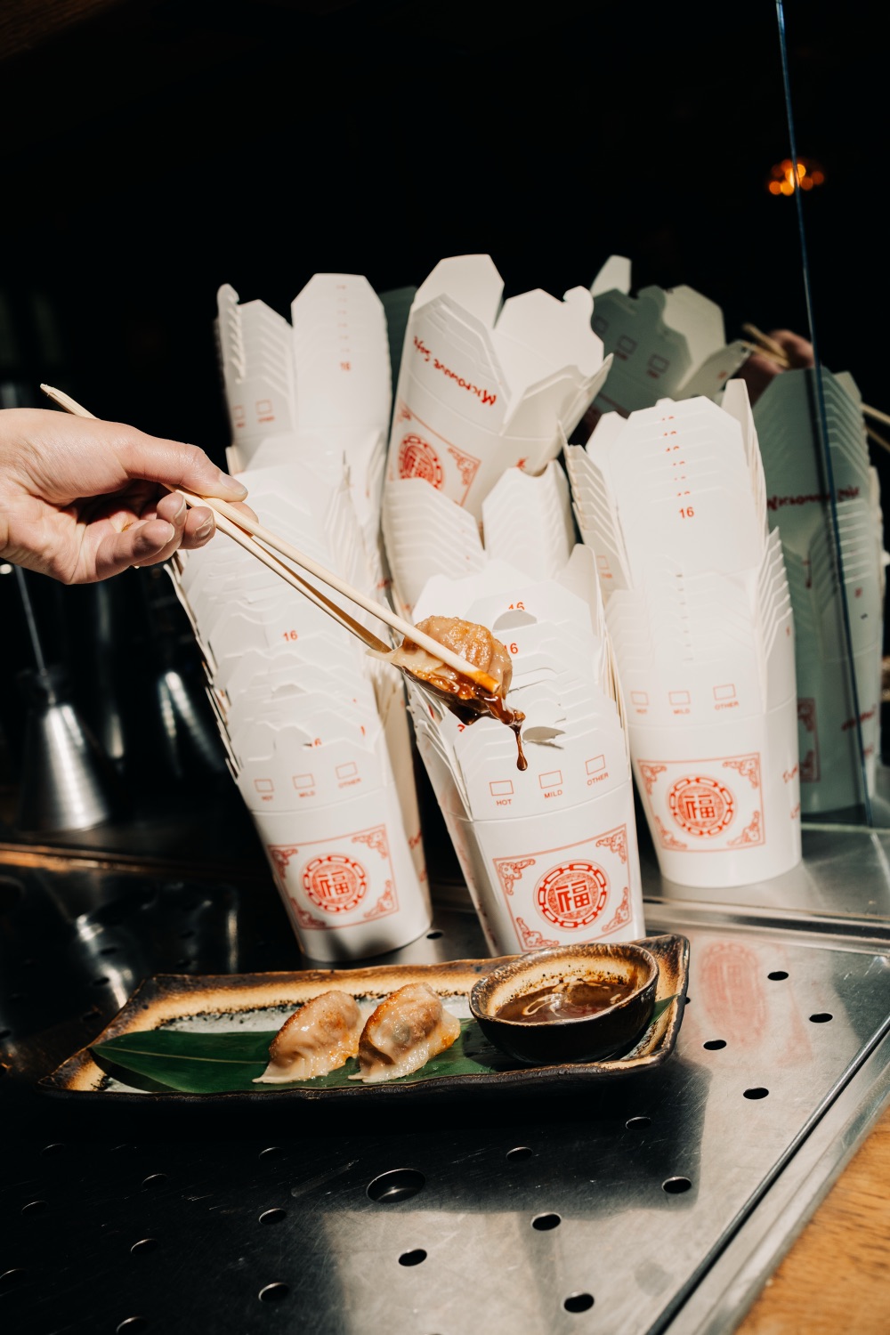 Chopsticks lifting a dumpling dripping with sauce, with takeout boxes in the background