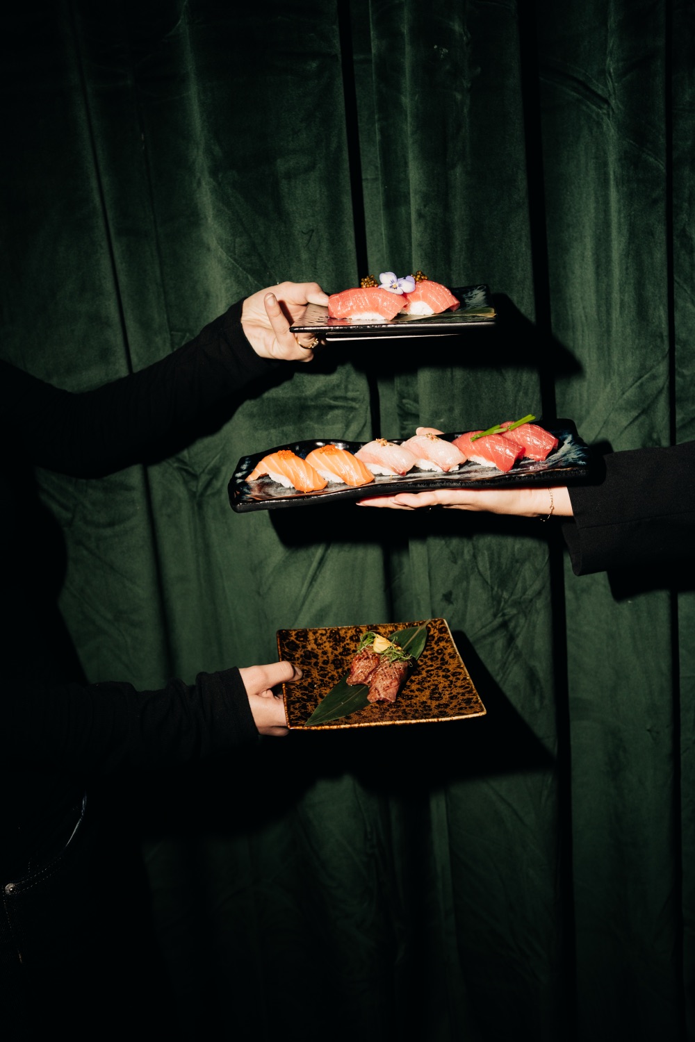 Three plates of sushi and wagyu held up against a green velvet curtain