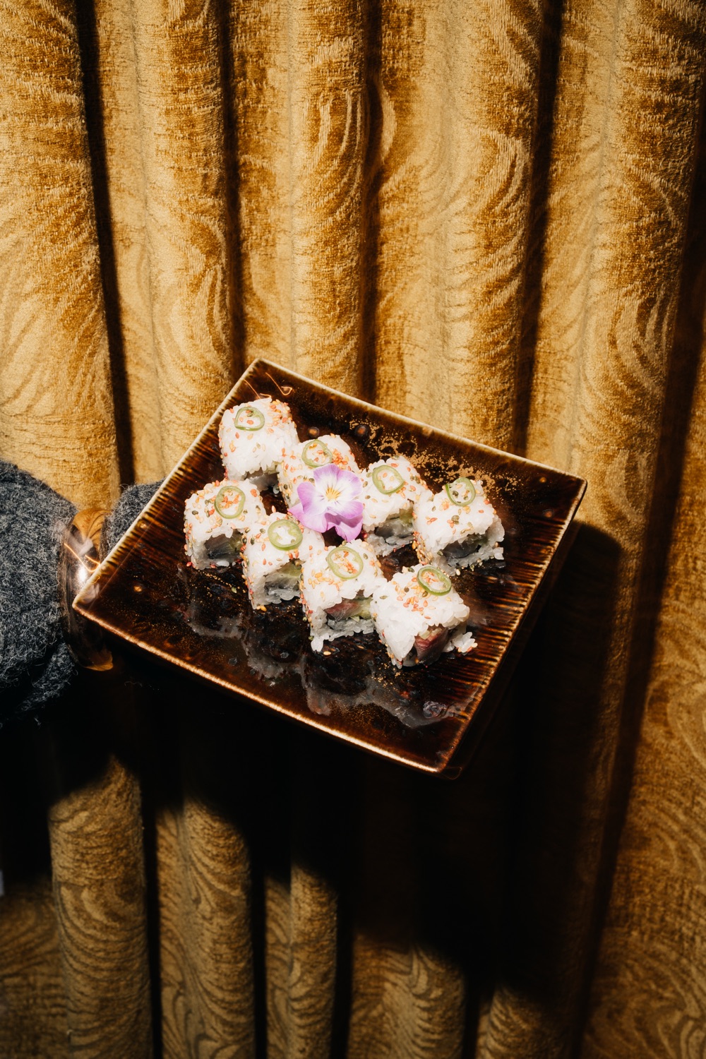 Sushi rolls topped with jalapeño slices and a purple flower against a gold curtain