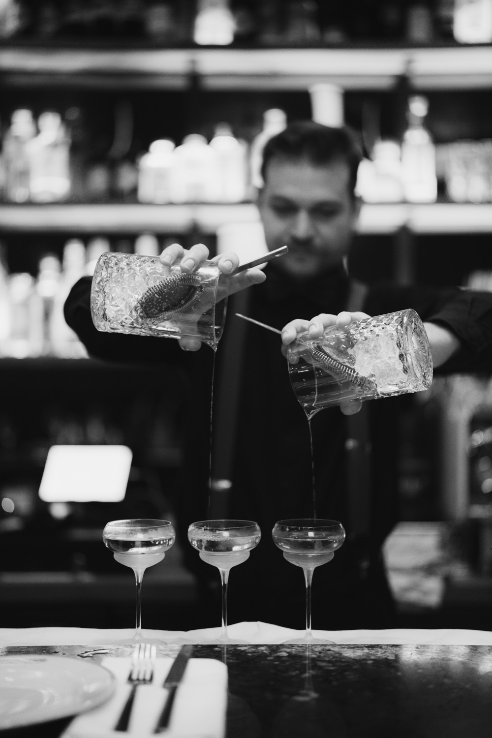 Black and white photo of a bartender pouring cocktails into three coupe glasses