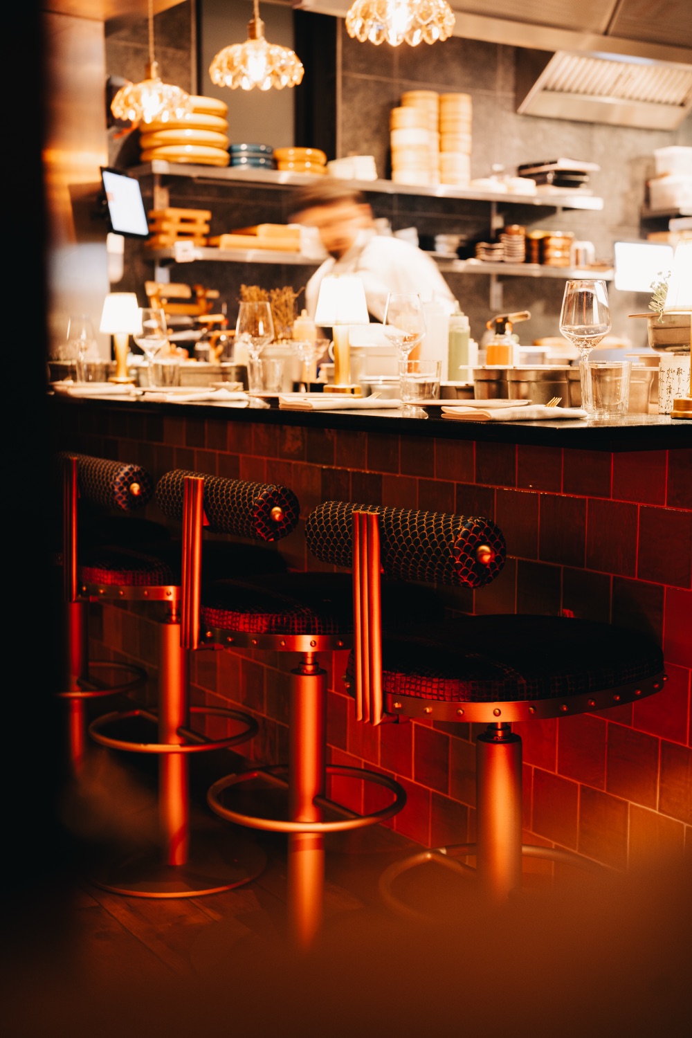 Warmly lit bar counter with stools and a chef working in the background